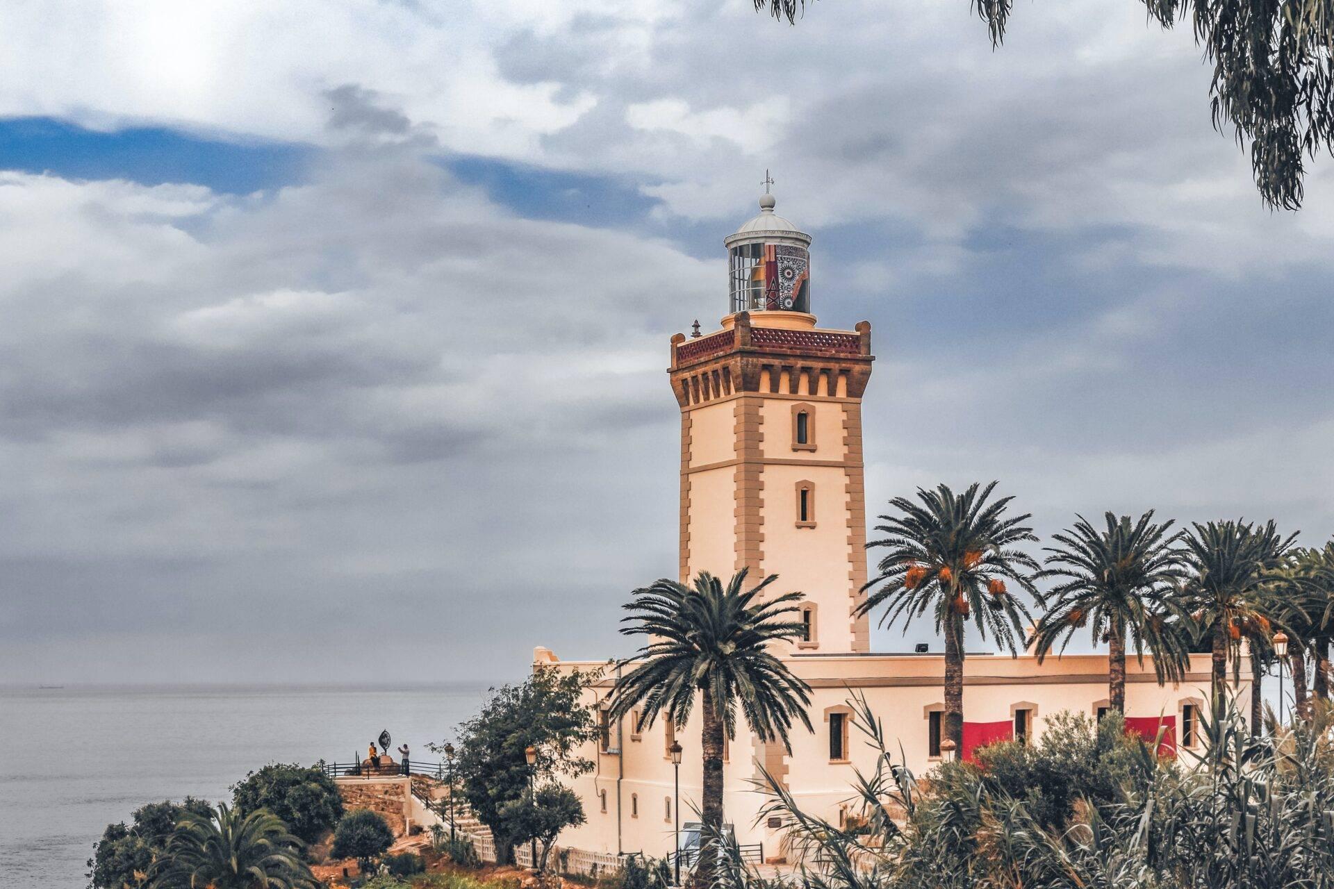 Un phare blanc côtier avec tour lanterneau, entouré de palmiers et surplombant la mer méditerranéenne sous un ciel nuageux