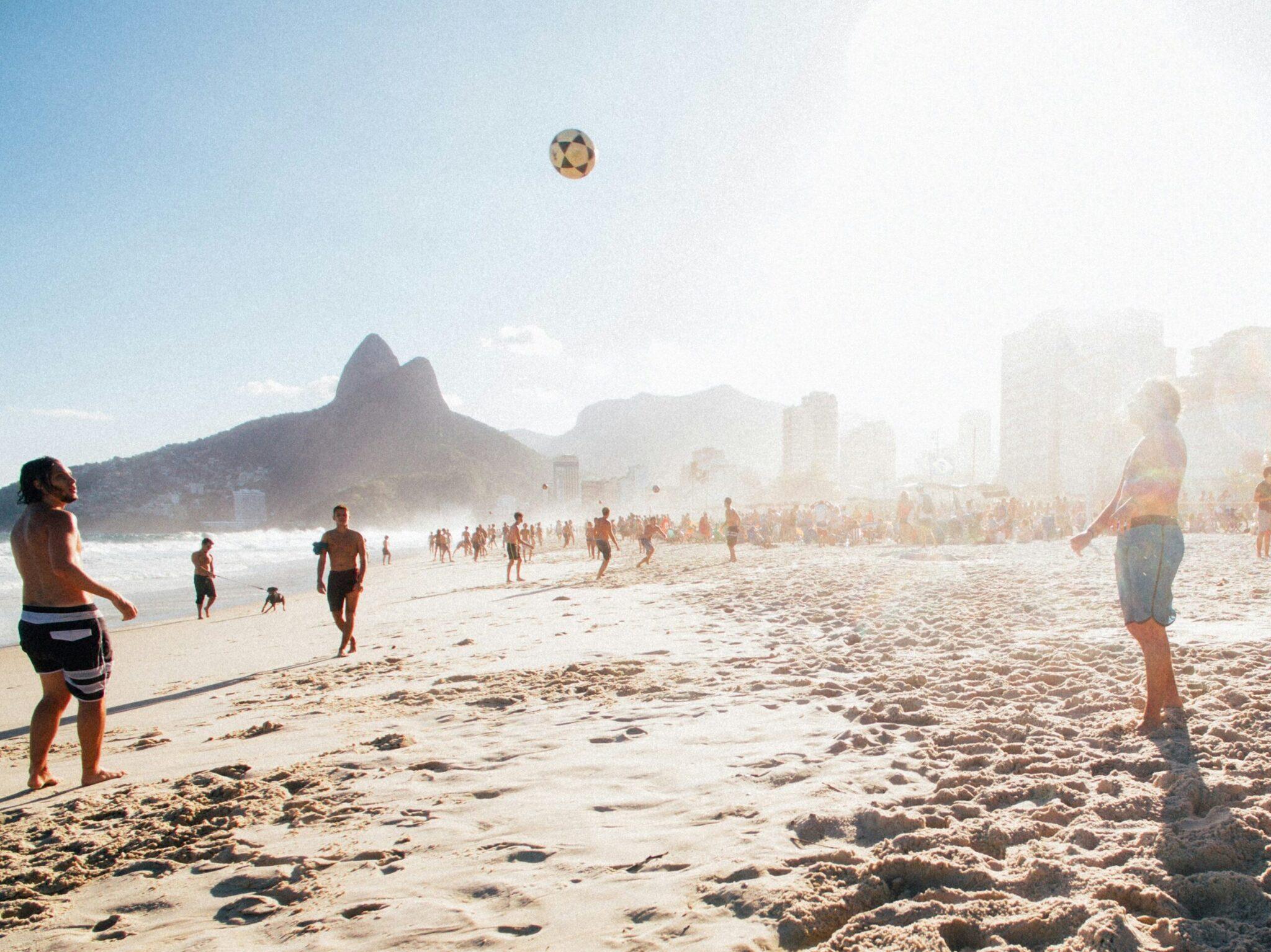 Groupe de personnes jouant au ballon sur une plage de sable fin avec montagne caractéristique et immeubles en arrière-plan