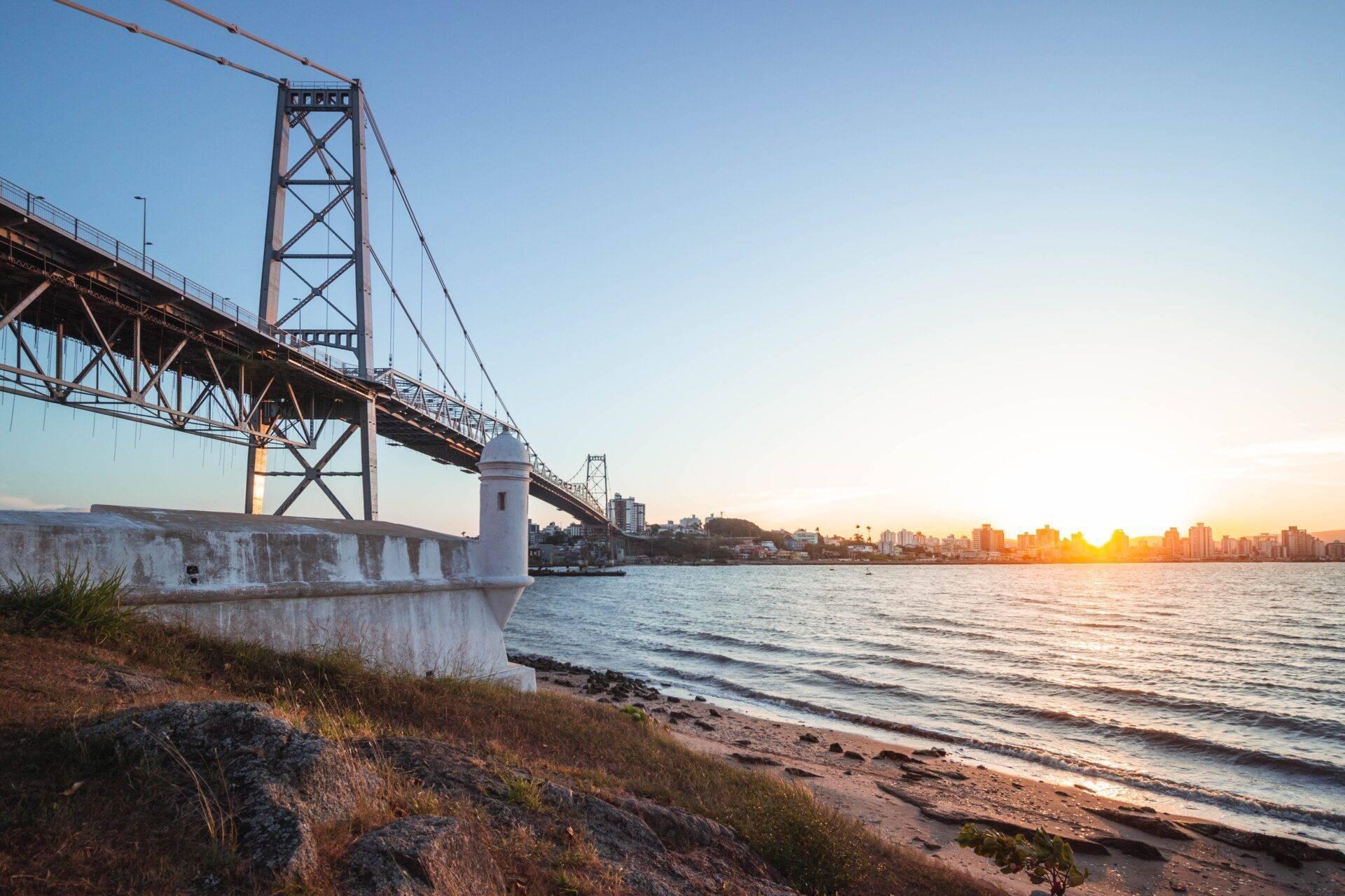 Un grand pont suspendu en acier rouille enjambe une baie au coucher de soleil, avec une ville côtière en arrière-plan