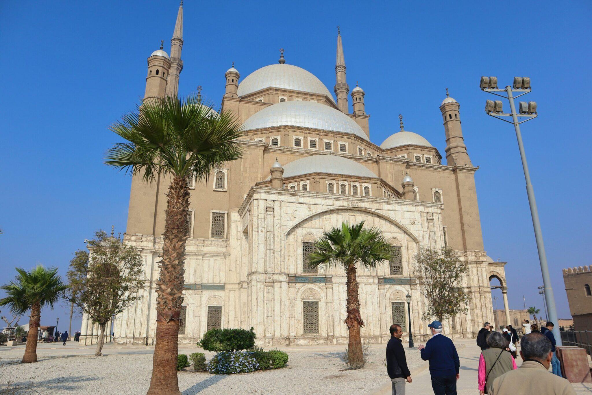 Une mosquée blanche au dôme imposant avec minarets, entourée de palmiers, sous un ciel dégagé