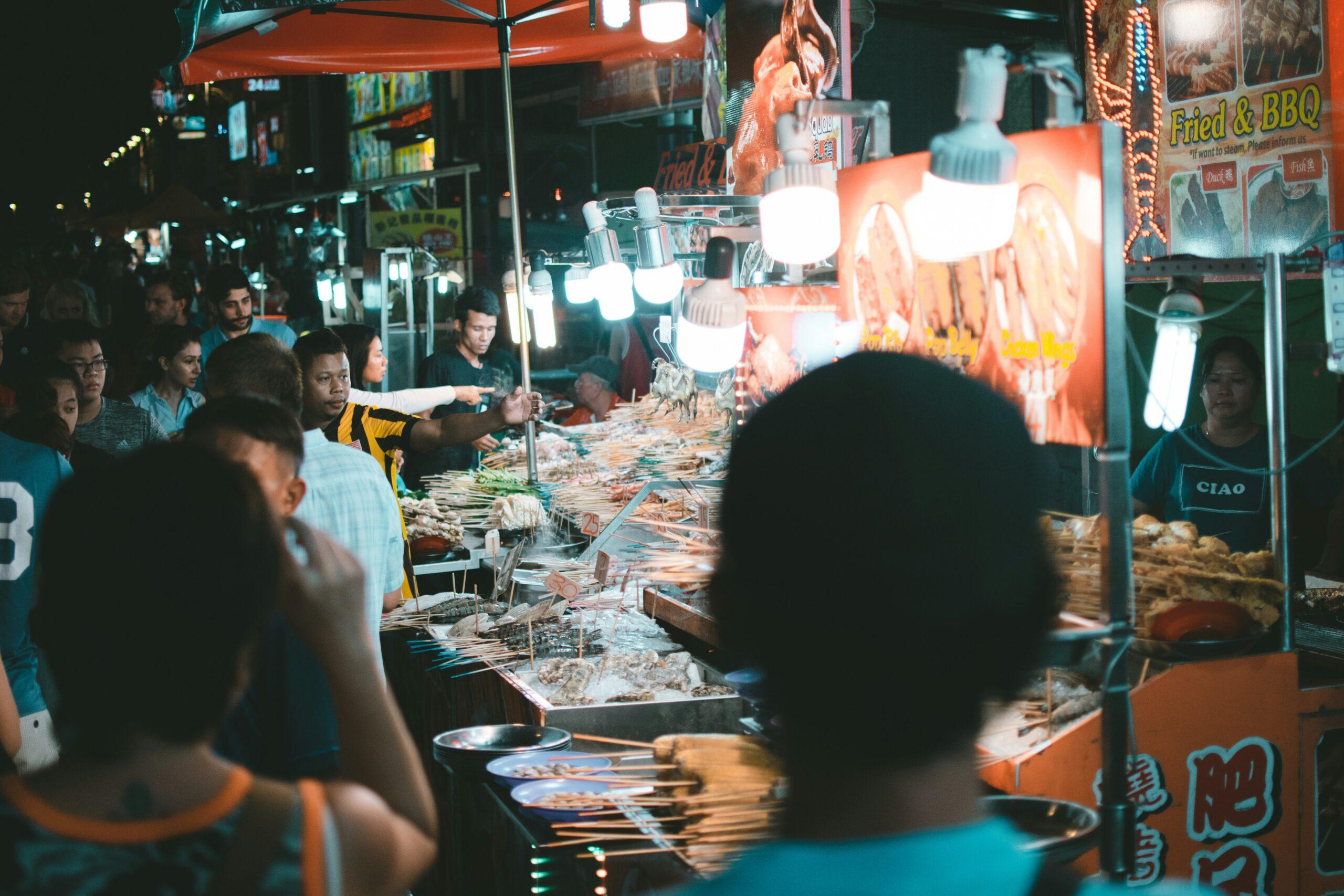 Un marché de rue nocturne avec des étals de nourriture grillée animé par des clients qui se pressent sous les lumières