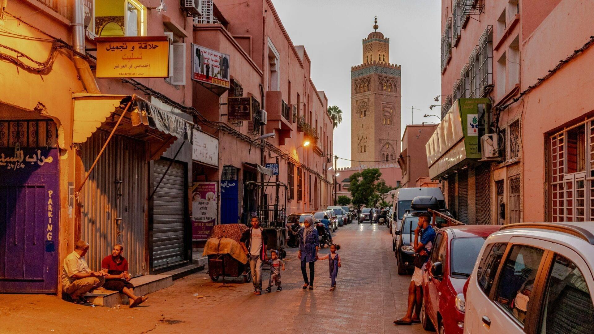 Une rue commerçante animée d’une médina marocaine avec une mosquée en arrière-plan, piétons et voitures