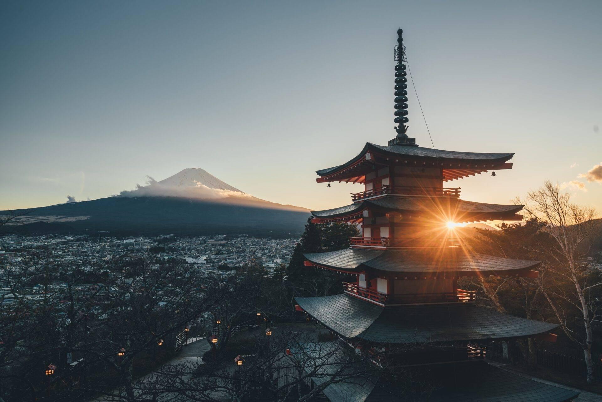 Un temple bouddhiste traditionnel au coucher de soleil avec le Mont-Fuji enneigé en arrière-plan et une ville illuminée