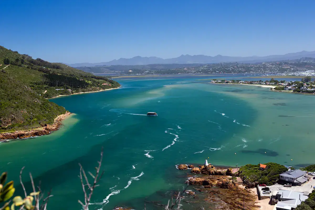 Vue aérienne d’une baie côtière aux eaux turquoise avec petit phare, montagnes en arrière-plan et habitations éparpillées
