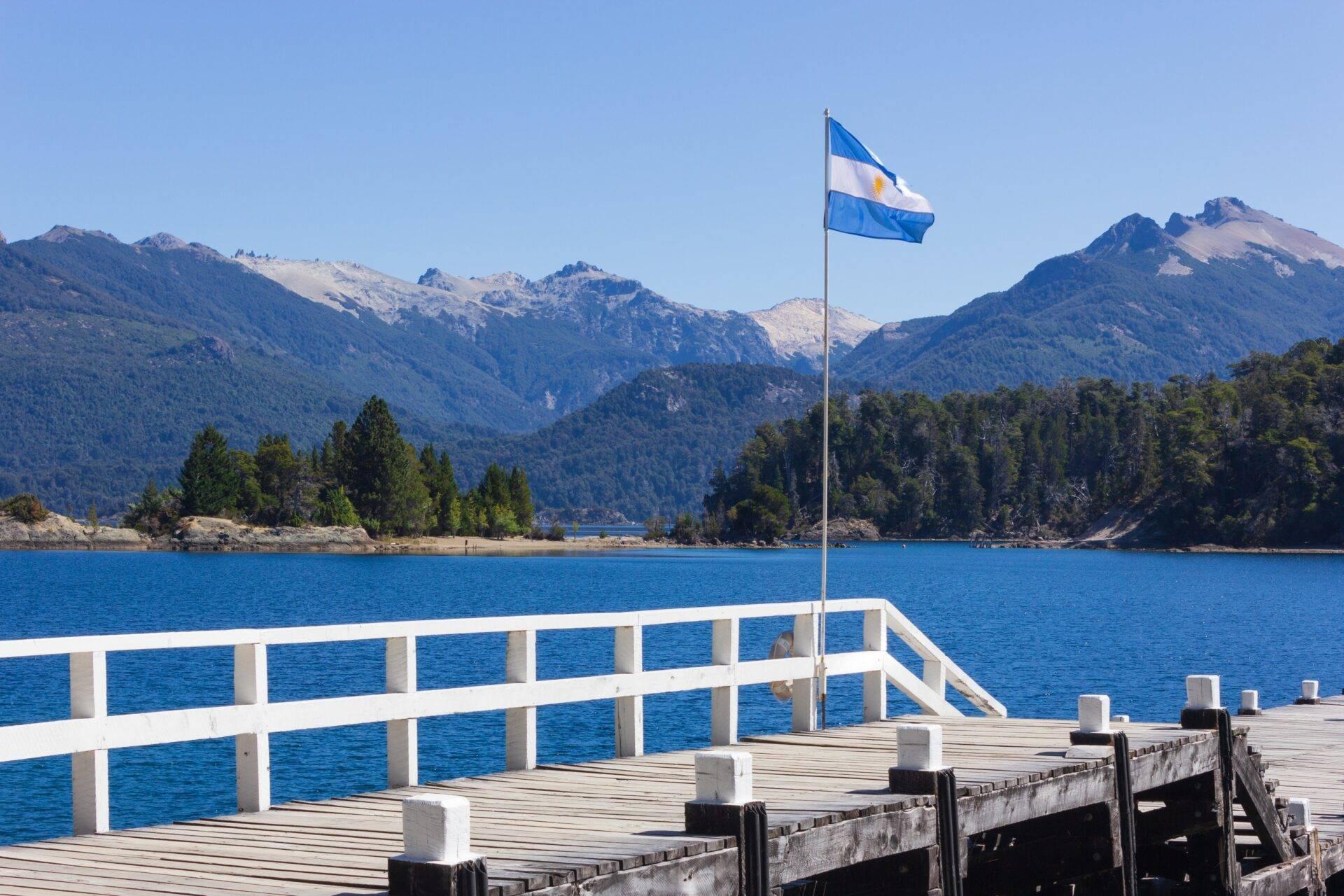 Un débarcadère en bois avec drapeau argentin face à un lac turquoise entouré de montagnes boisées