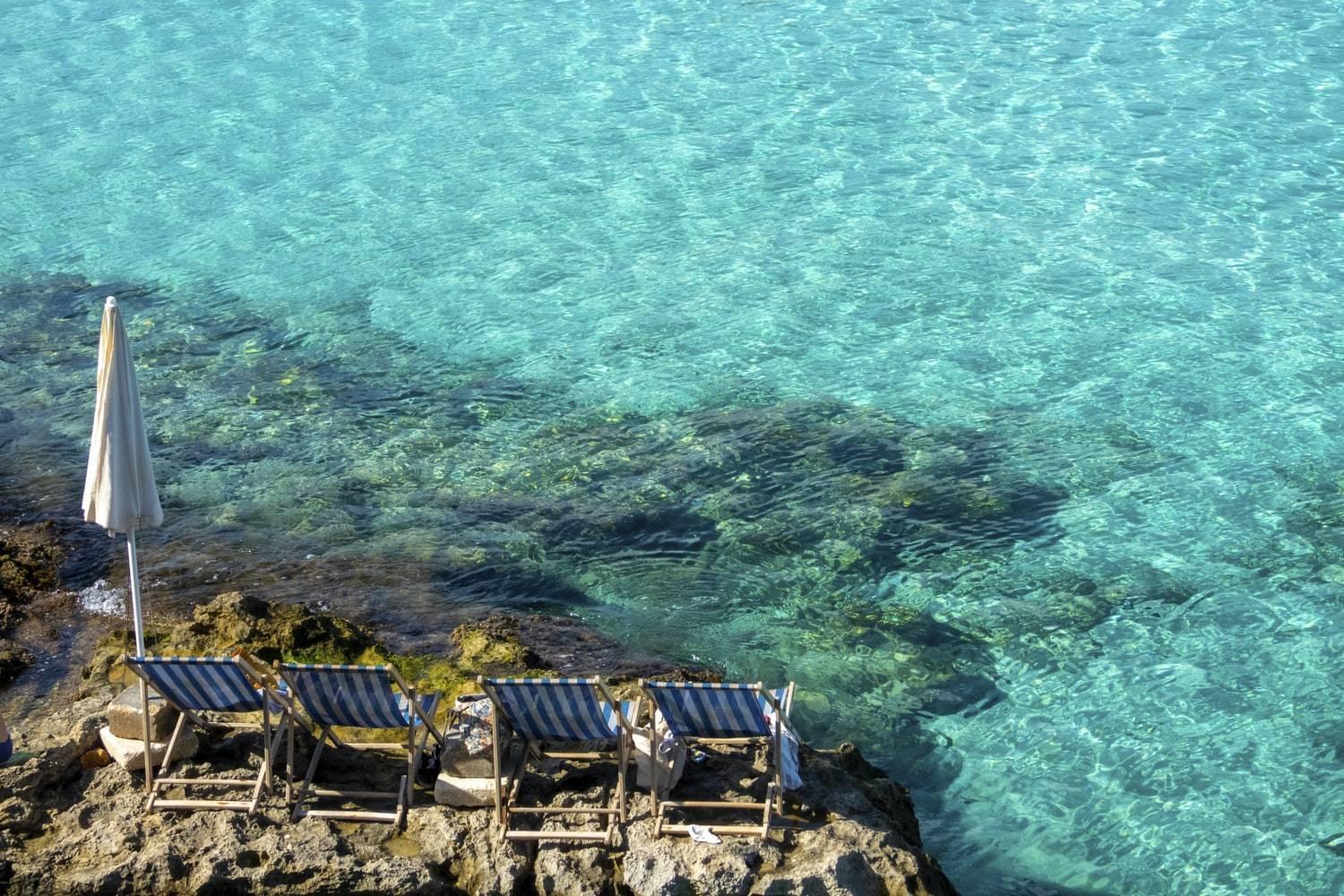 Chaises longues rayées et parasol blanc sur des rochers face à une mer turquoise cristalline méditerranéenne