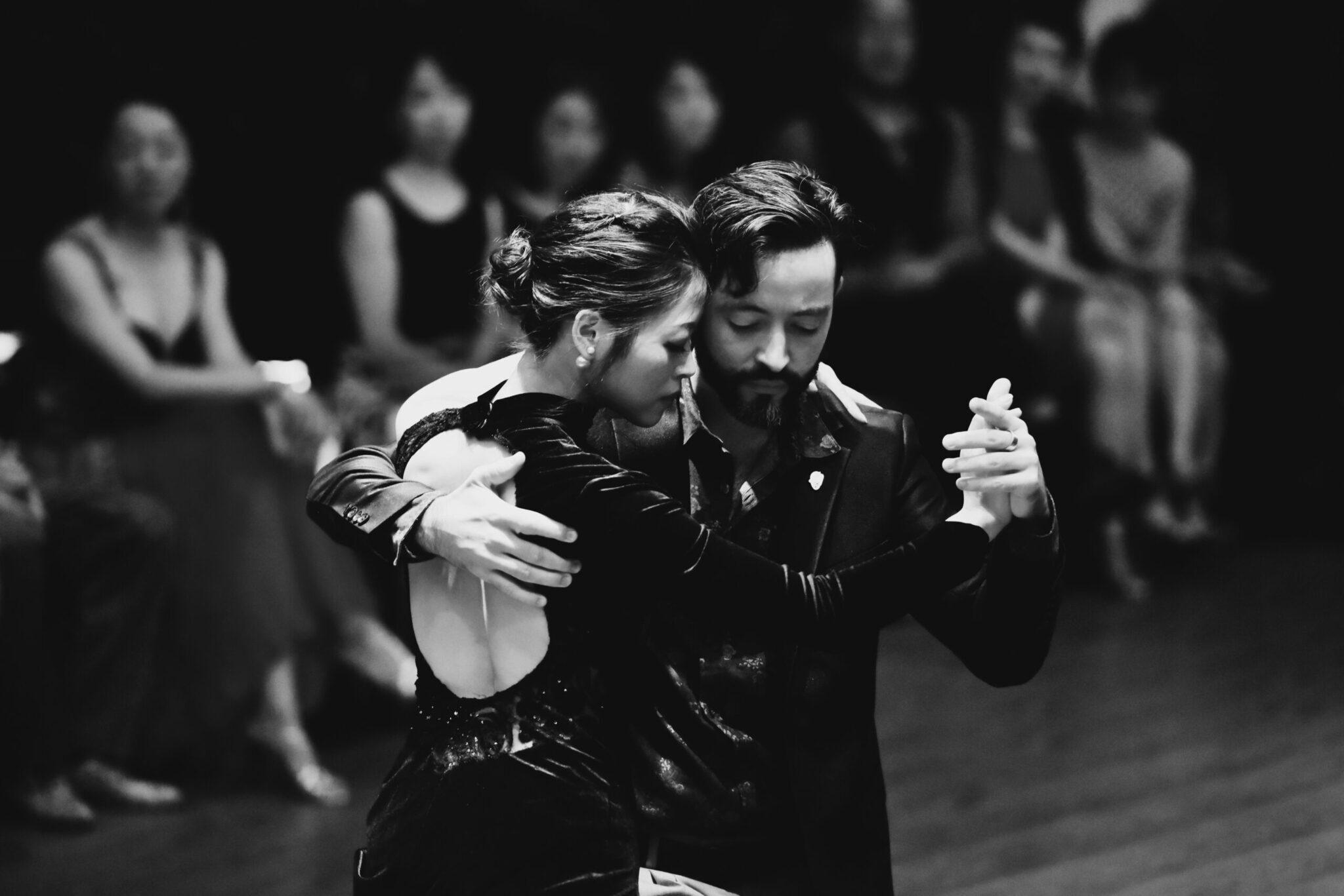 Couple dansant le tango en costume noir devant des spectateurs dans une ambiance théâtrale en noir et blanc