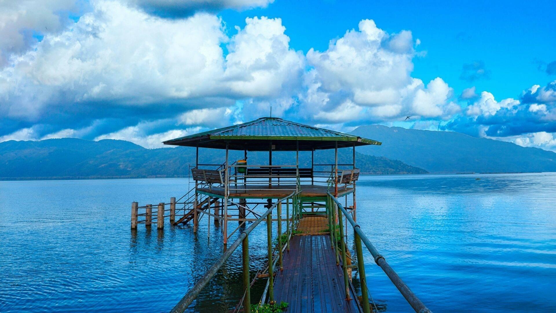 Un ponton en bois avec abri ombragé s’étend sur un lac turquoise, montagnes boisées en arrière-plan sous ciel nuageux