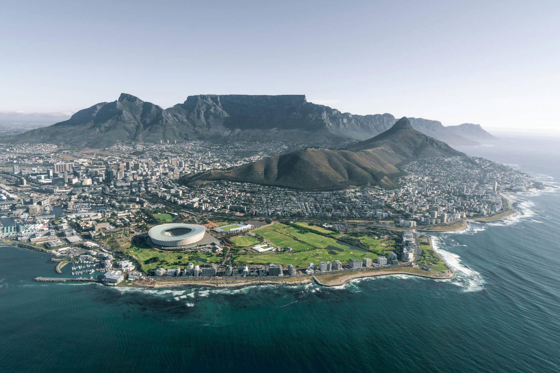 Vue aérienne d’une ville côtière avec un stade circulaire, montagne majeure en arrière-plan et plage verdoyante en bord de mer