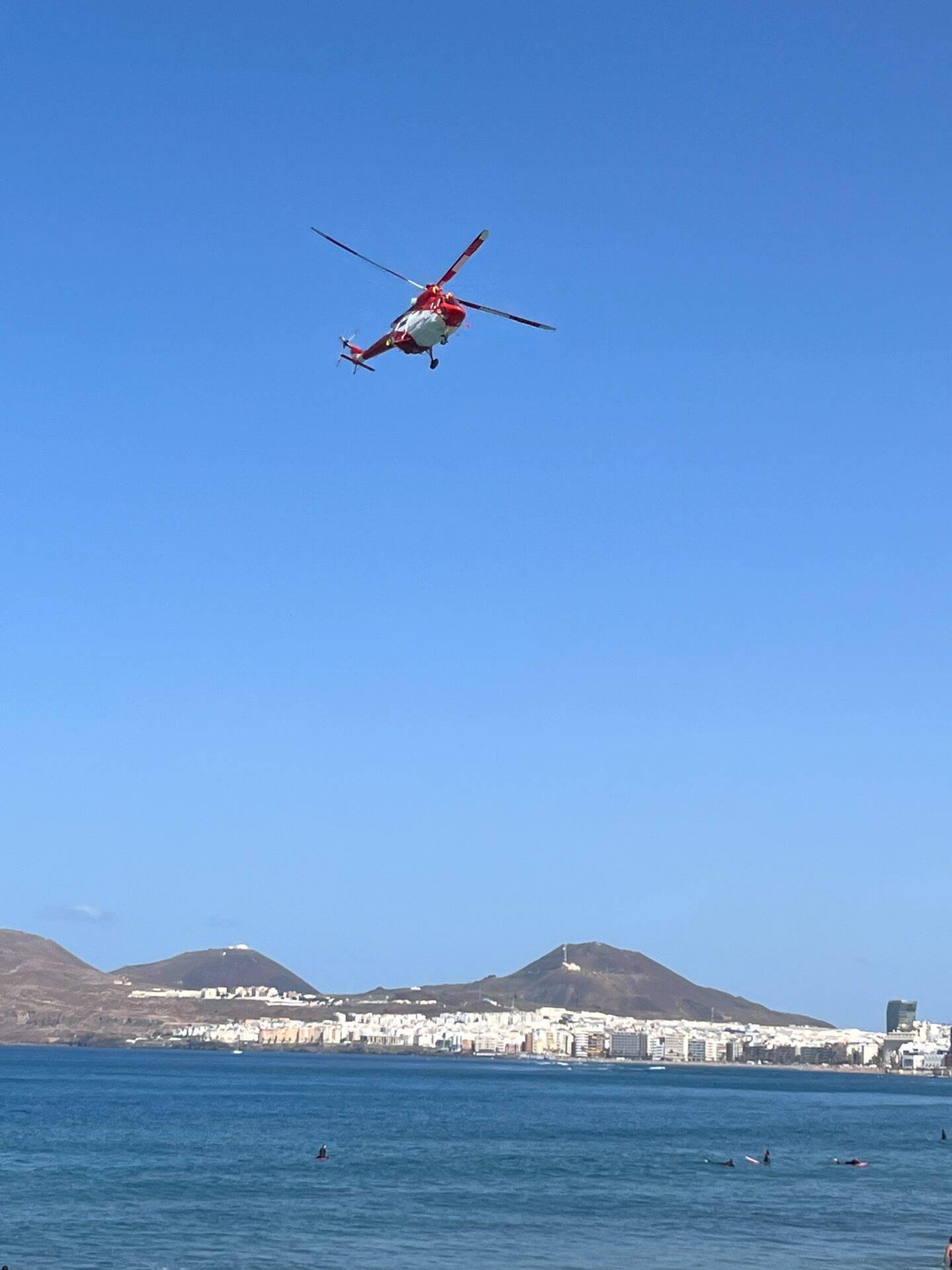 Un hélicoptère de secours survole une plage avec des baigneurs face à une ville côtière et des volcans