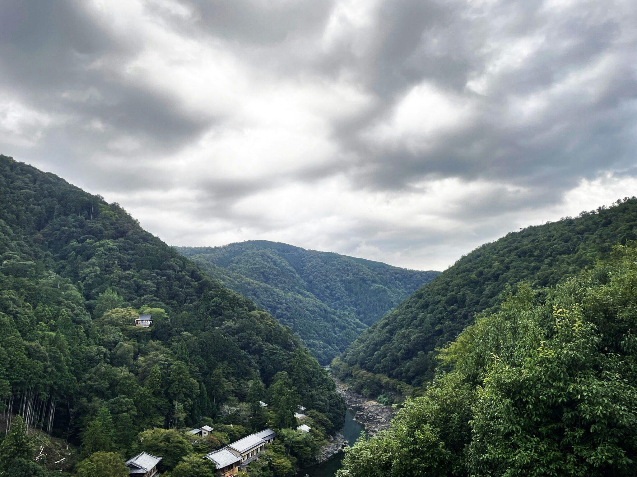 Une vallée boisée avec rivière, maisons traditionnelles et montagnes densément couvertes de végétation sous ciel nuageux