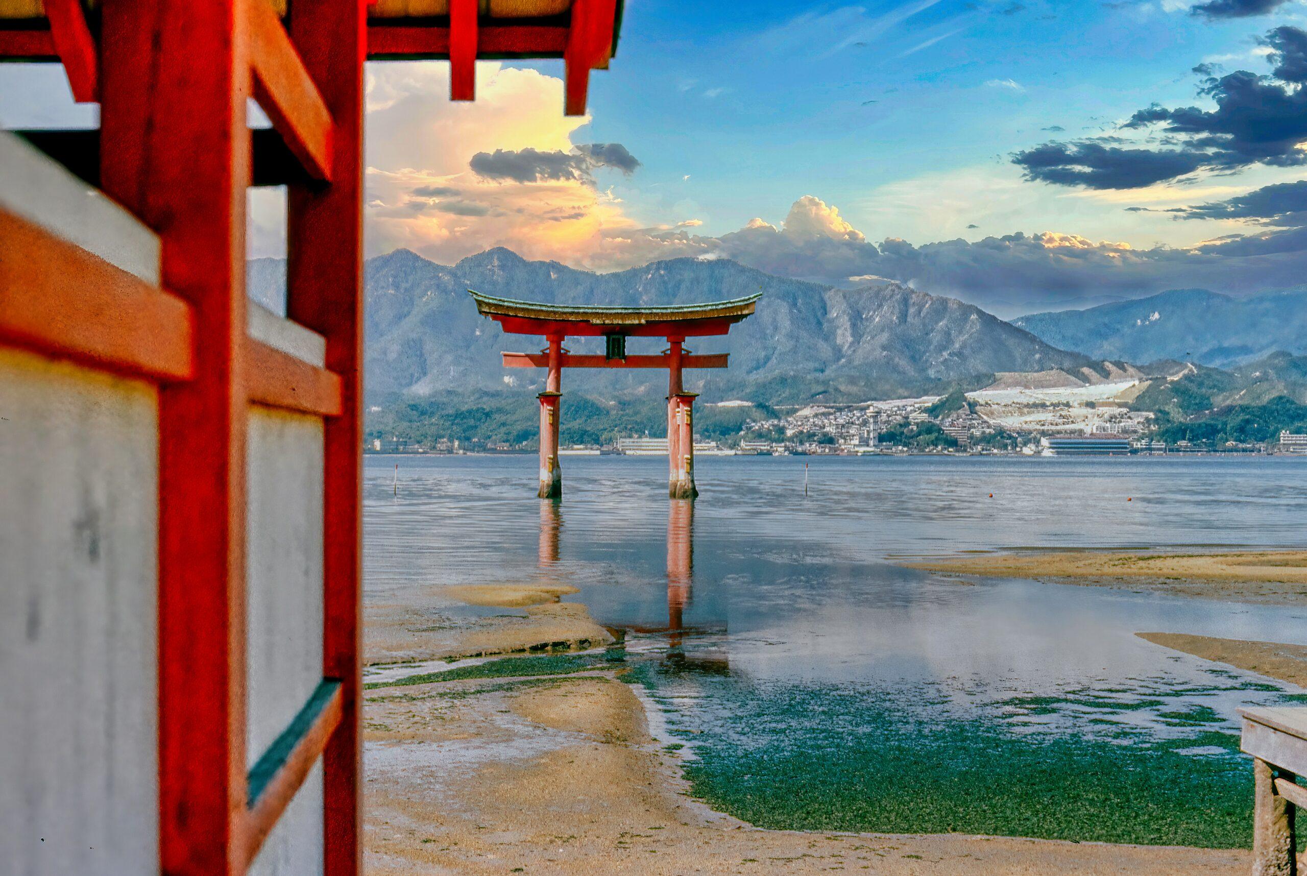 Un torii rouge traditionnel émerge des eaux peu profondes face à des montagnes, avec un temple en arrière-plan