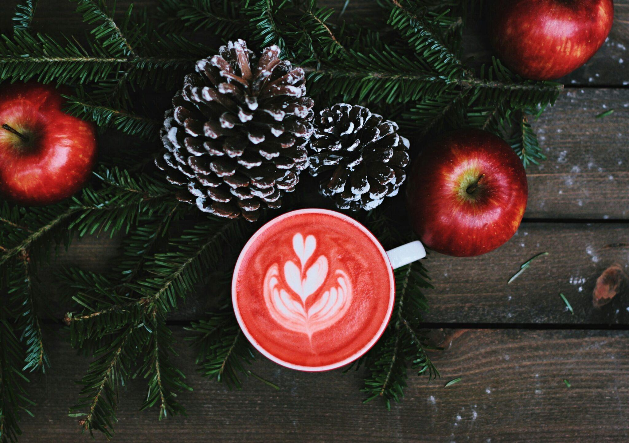 Tasse de café avec latte art en forme de cœur entourée de branches de sapin, pommes rouges et pommes de pin enneigées