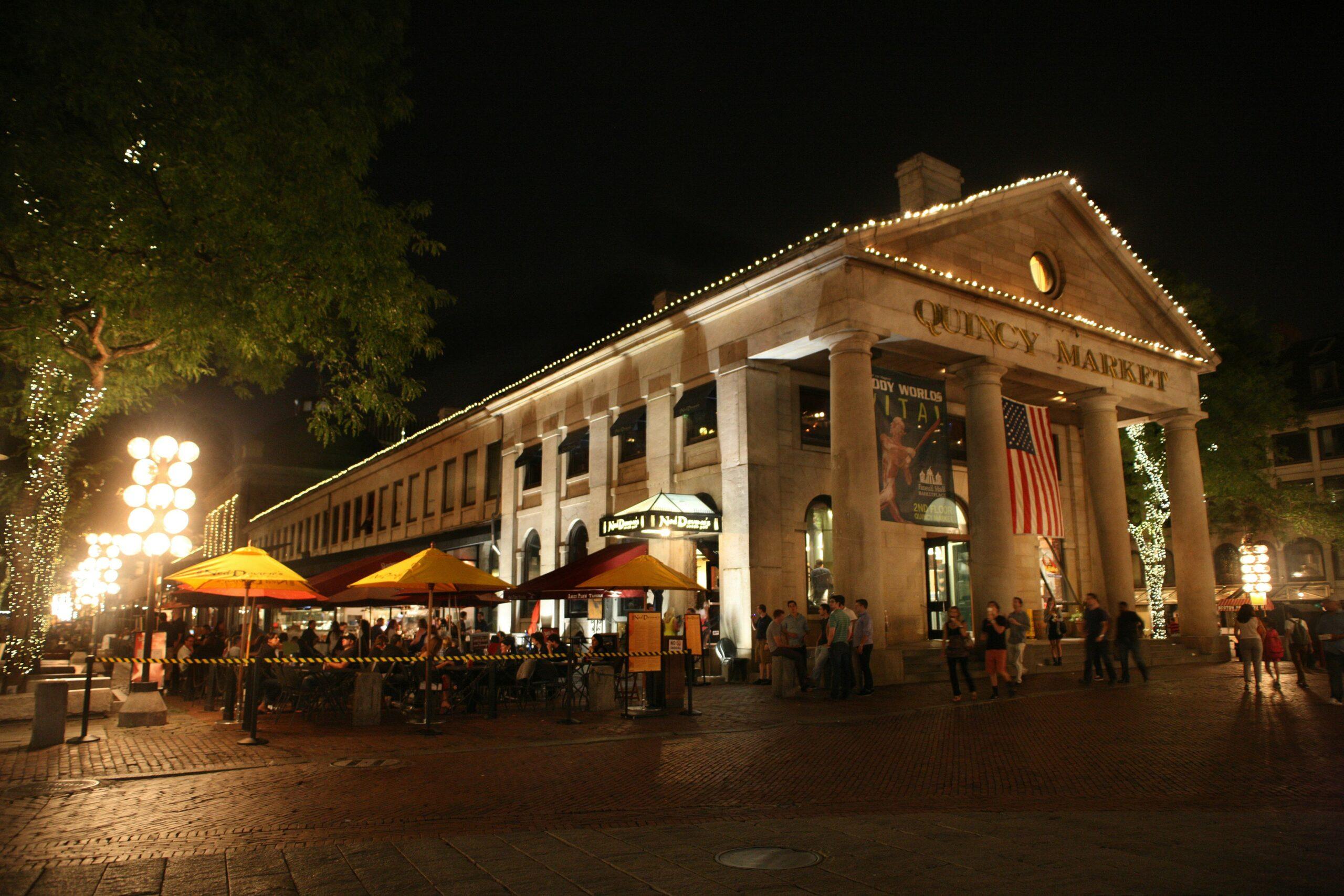 Façade illuminée d’un marché historique de style néoclassique avec colonnes, terrasse sous parasols et passants en soirée