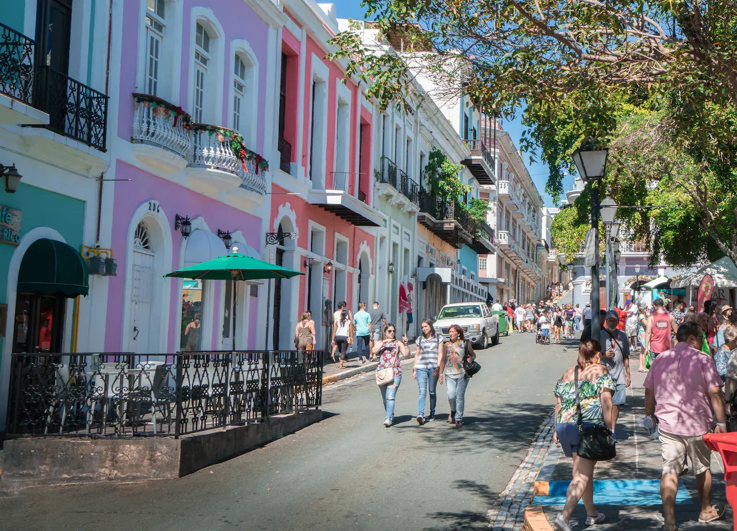 Une rue piétonne colorée bordée de bâtiments pastel rose et turquoise avec des touristes flânant sous le soleil