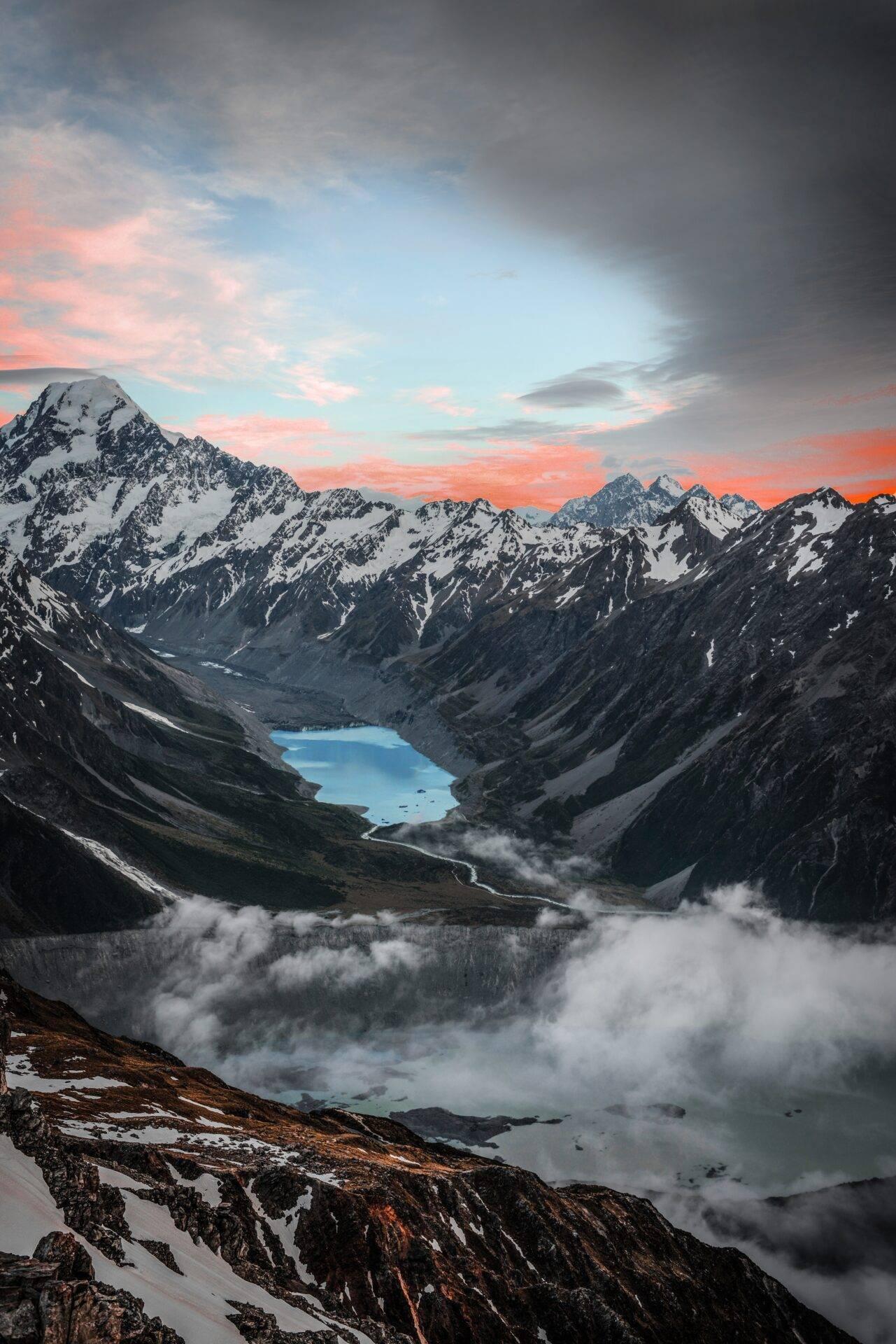 Chaîne montagneuse enneigée au coucher de soleil avec lac turquoise encaissé, vallée brumeux et ciel rose