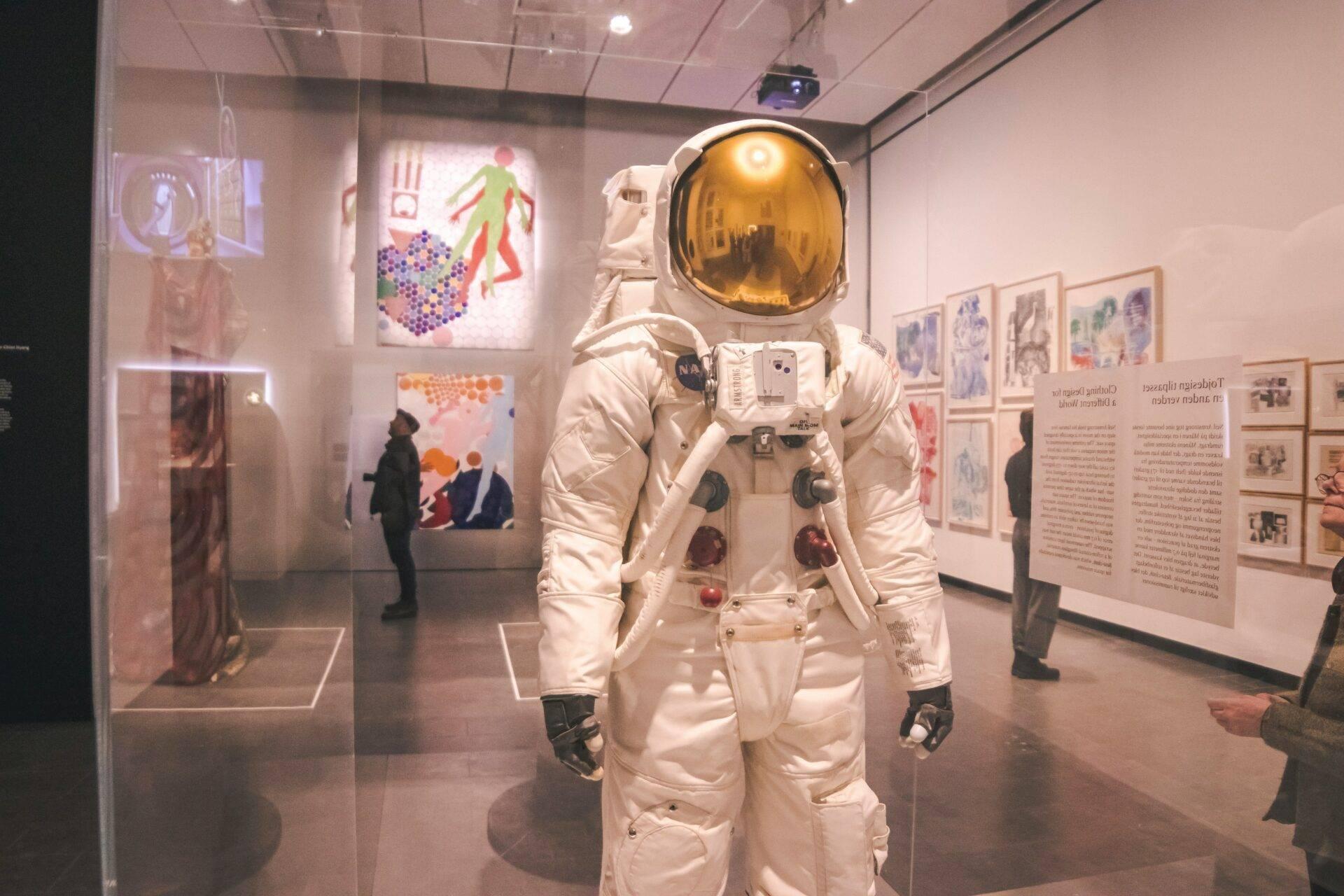 Un combinaison spatiale blanche avec casque doré exposée dans un musée, visiteurs observant l’exposition en arrière-plan