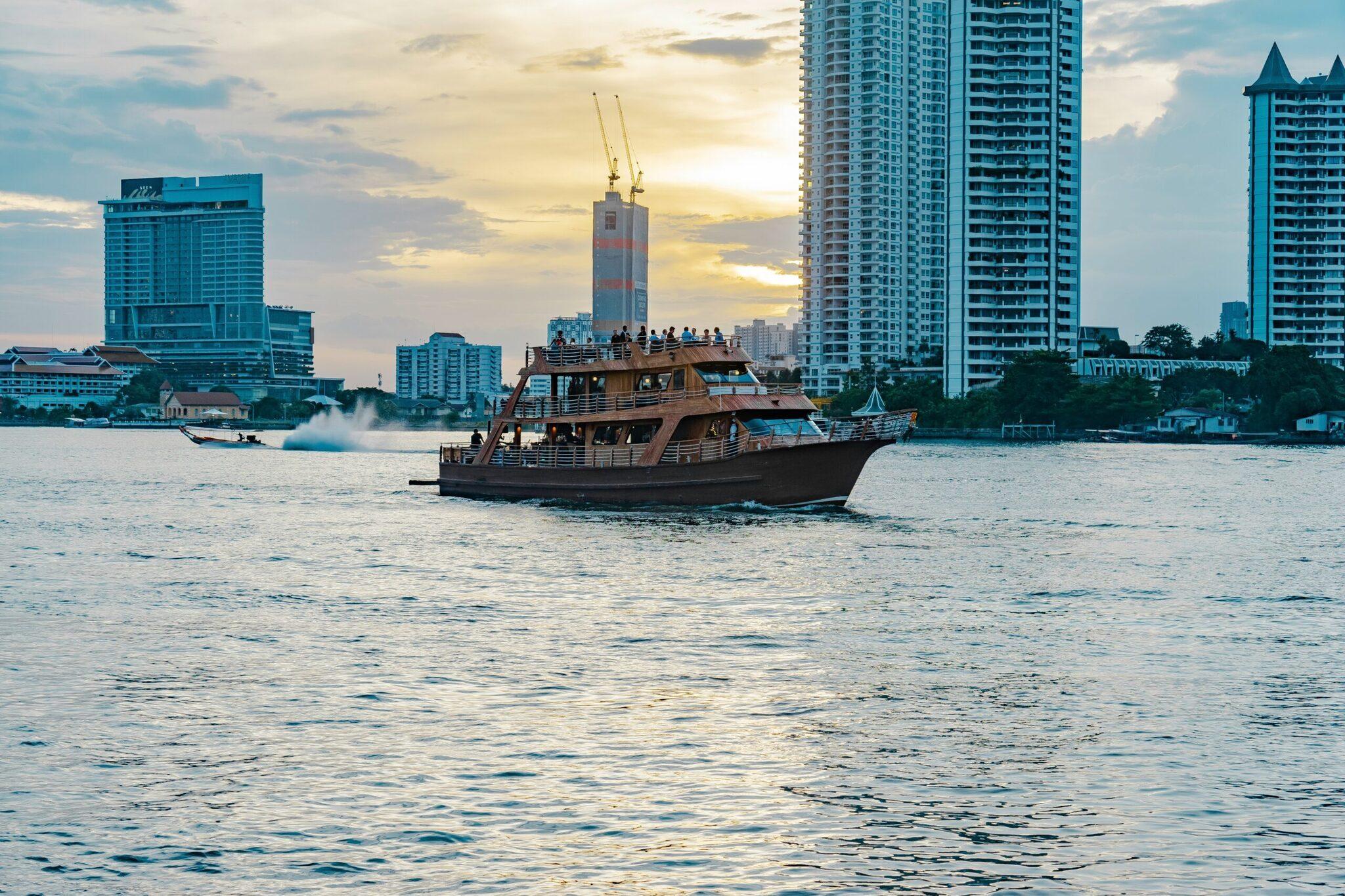 Bateau touristique traditionnel naviguant sur un fleuve avec gratte-ciel et temple en arrière-plan au coucher du soleil
