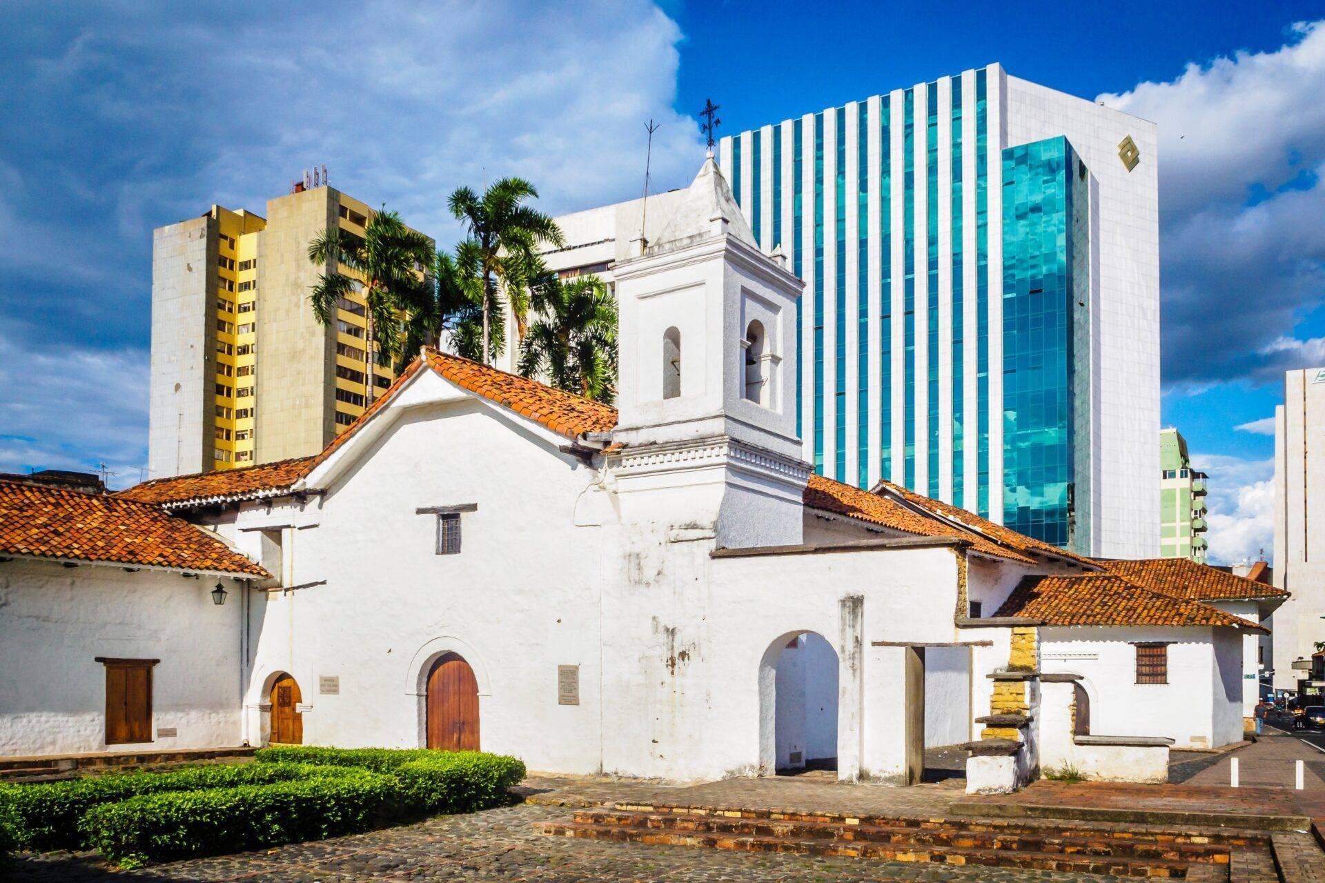 Une église coloniale blanche aux toits de tuiles orangées contraste avec les bâtiments modernes en arrière-plan sous un ciel bleu