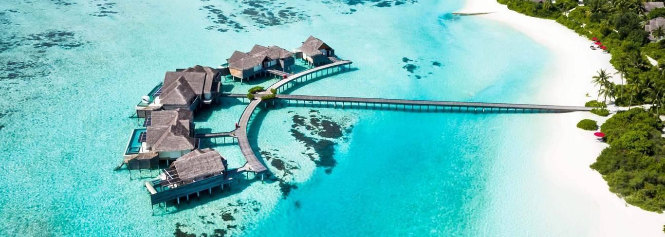 Vue aérienne d’un resort tropical avec bungalows sur pilotis, plage de sable blanc et lagon turquoise cristallin