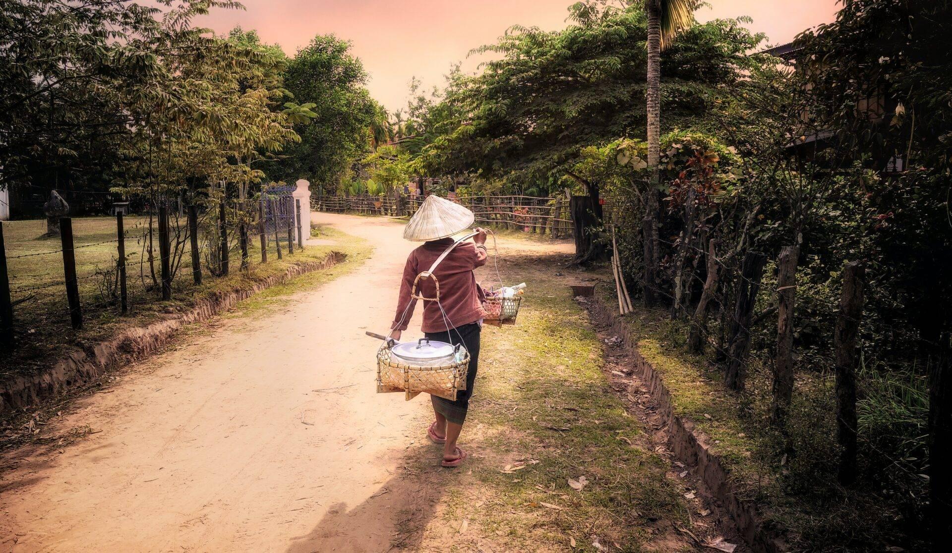 Une femme portant un chapeau conique marche sur un chemin de terre bordé de verdure en Asie du Sud-Est rurale