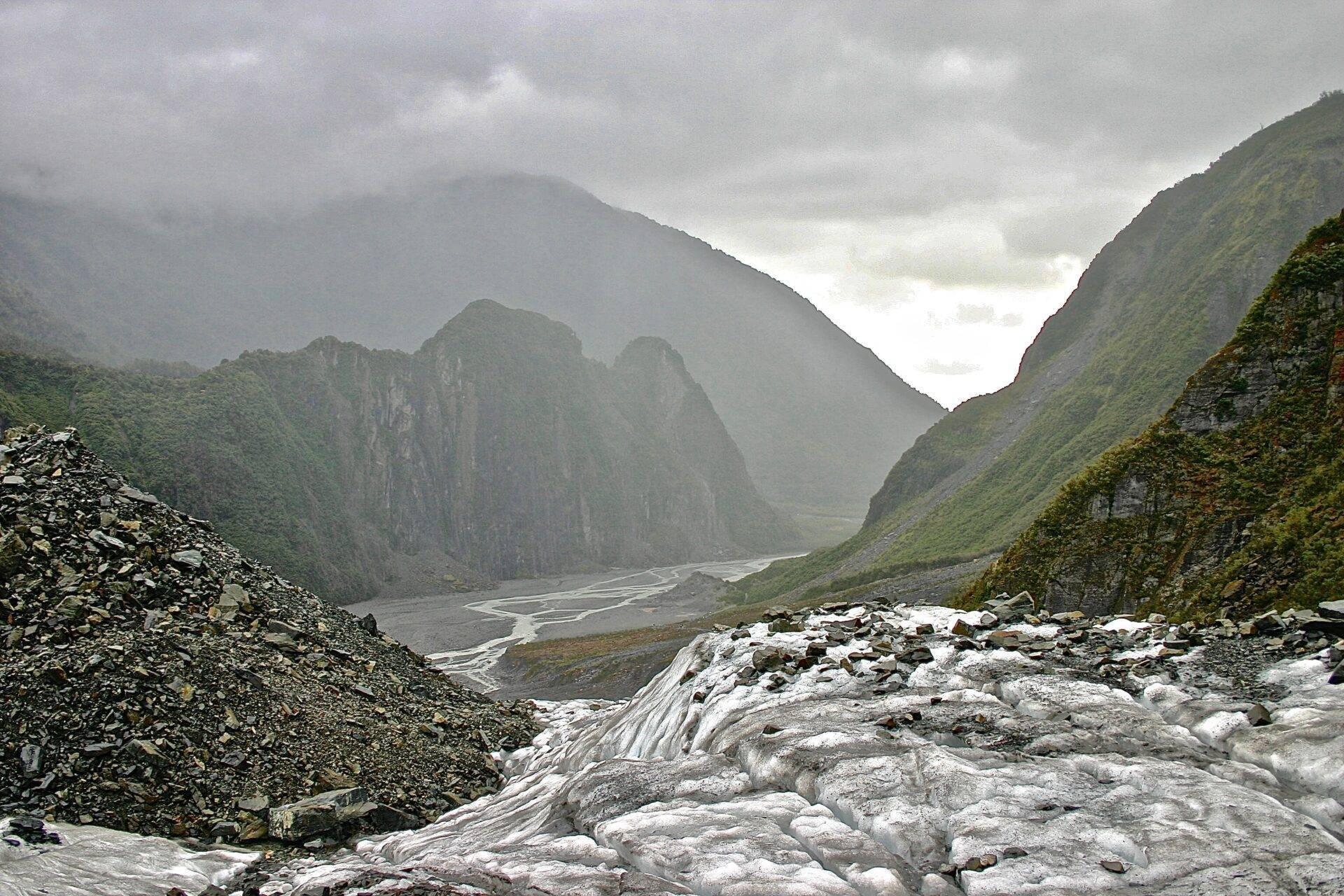 Vallée montagneuse avec glacier, rivière sinueuse et parois rocheuses escarpées sous ciel nuageux