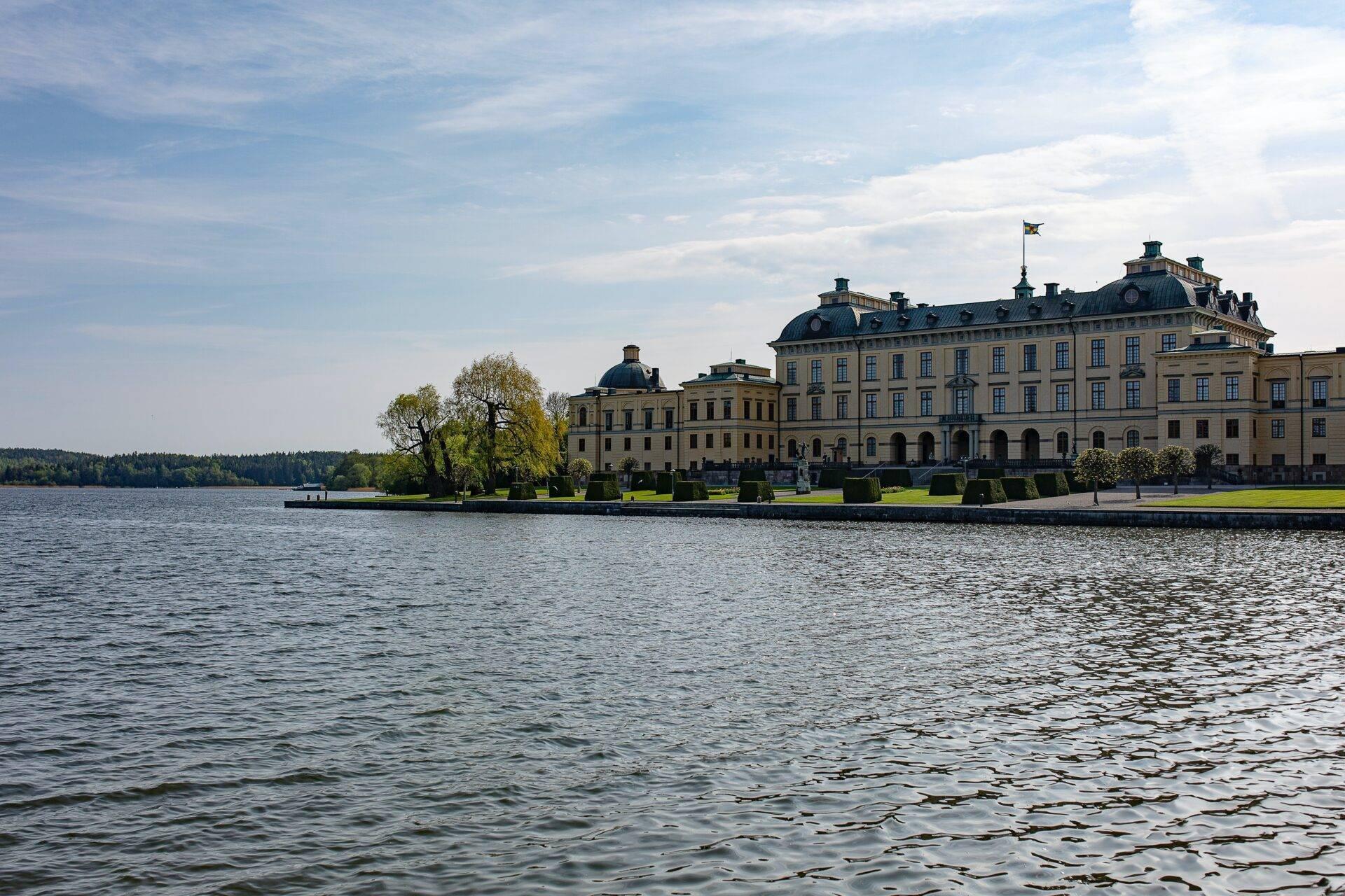 Un majestueux palais jaune de style baroque au bord d’un lac calme, entouré de verdure et de ciel nuageux