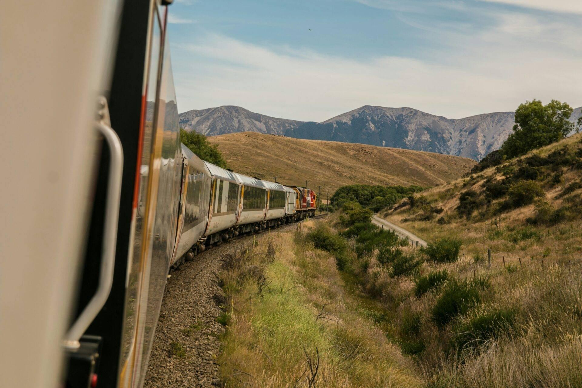 Un train de passagers traverse une vallée montagneuse aride avec des collines boisées sous un ciel dégagé