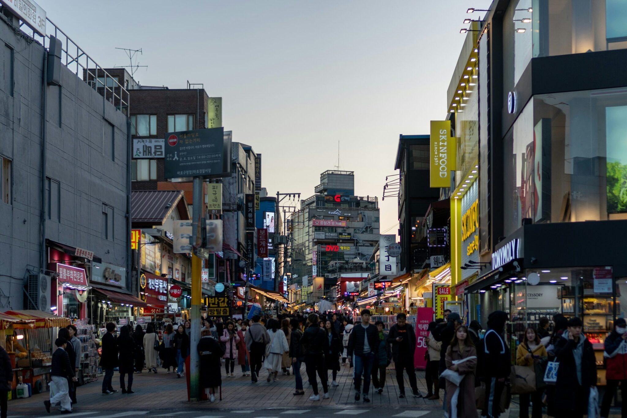 Une rue commerciale animée de Séoul avec foule, enseignes lumineuses coréennes et boutiques en début de soirée