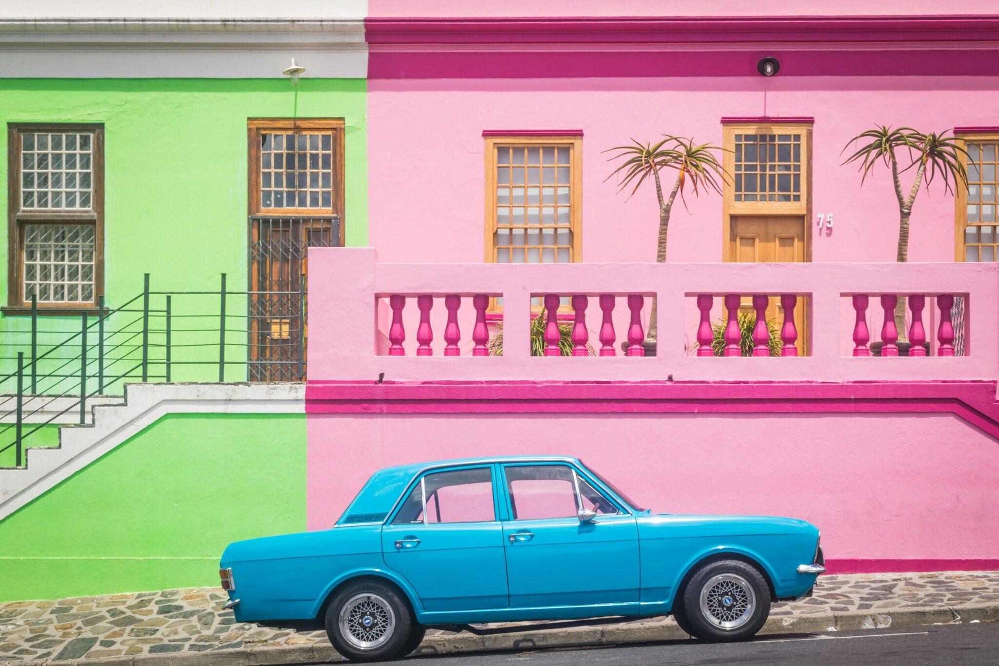 Une voiture bleue vintage garée devant des façades colorées rose et vert avec balcons ornementaux et palmiers