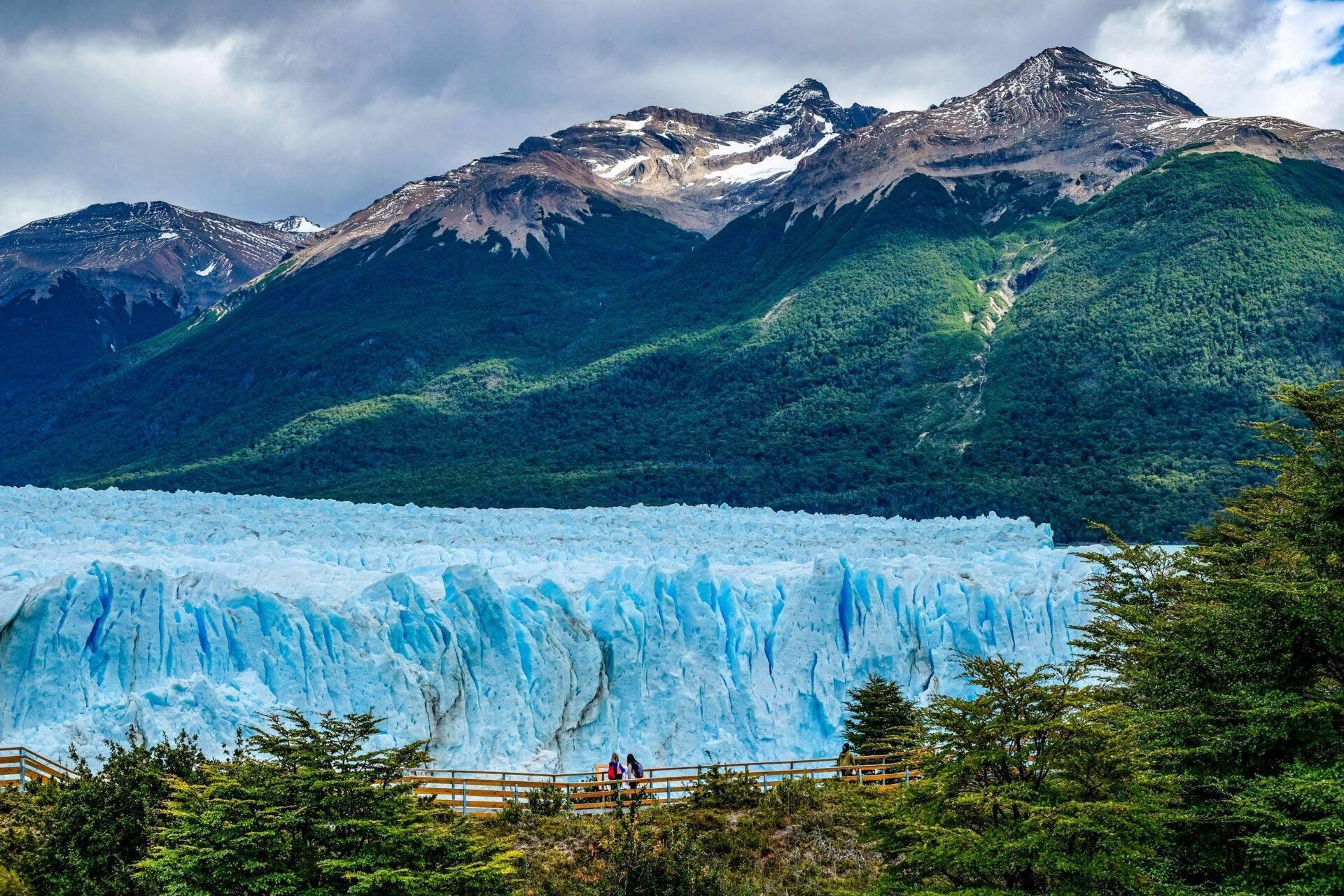 Un glacier bleu imposant face à des montagnes enneigées et une forêt dense sous un ciel nuageux