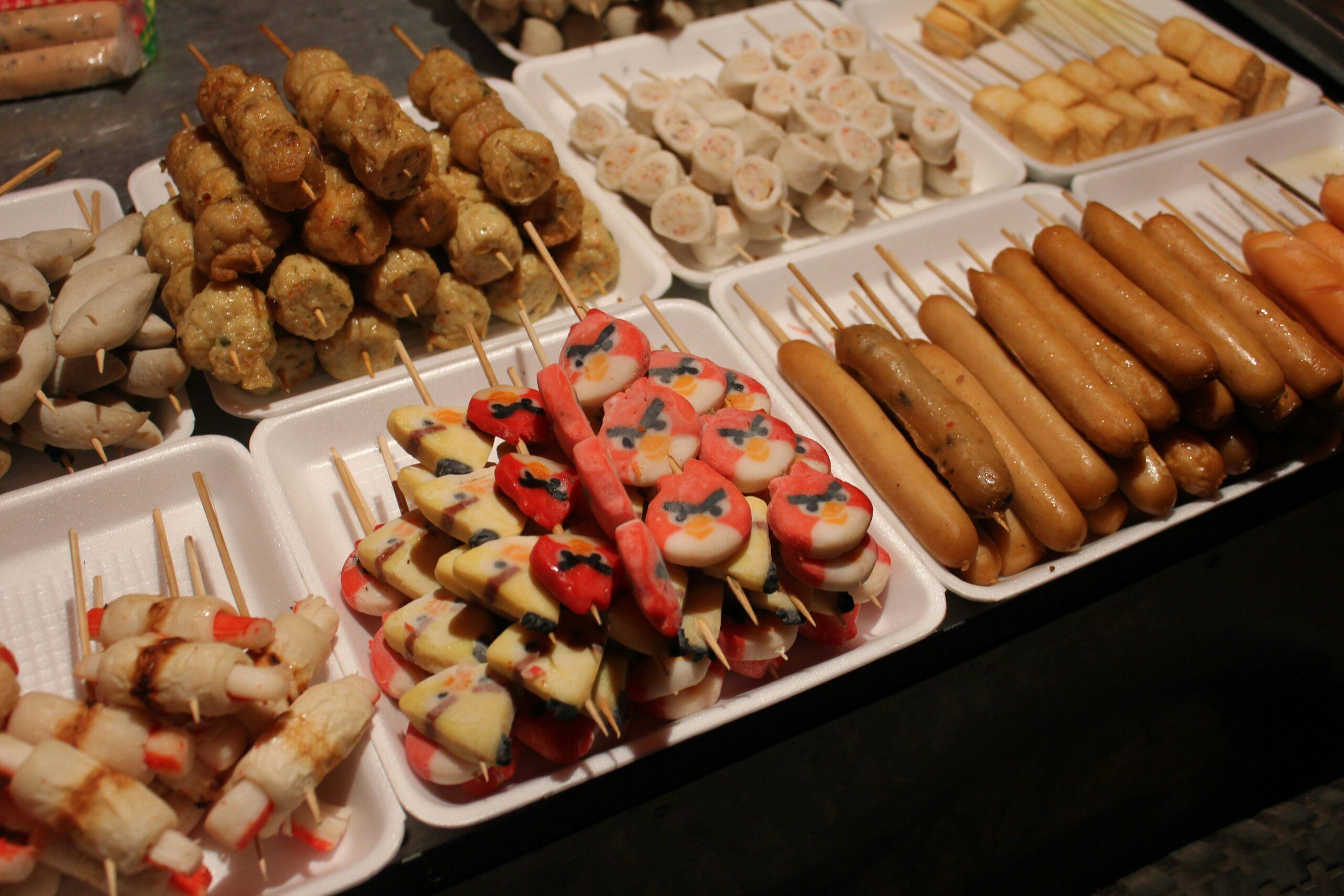 Étal de brochettes grillées et saucisses sur des plateaux blancs dans une rue commerçante asiatique