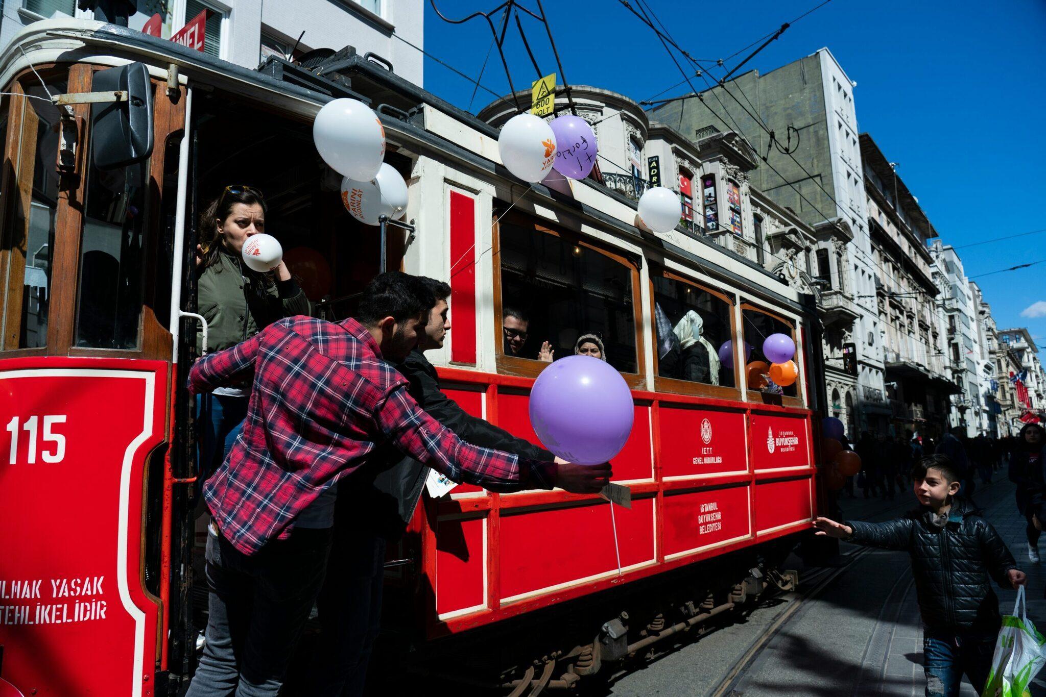 Un tramway rouge historique décoré de ballons blancs et violets circule dans une rue animée d’une ville
