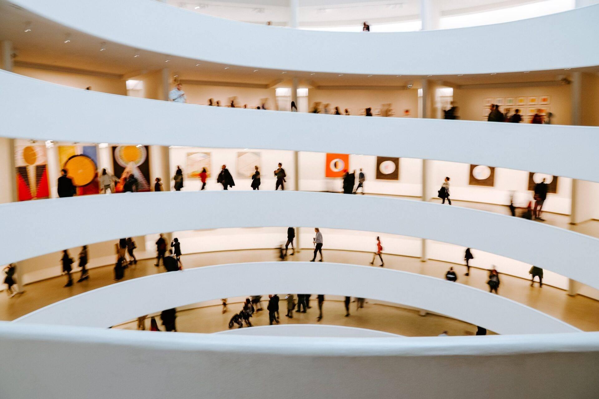 Vue en plongée d’un musée aux étages spiralés blancs avec visiteurs déambulant et œuvres d’art colorées exposées