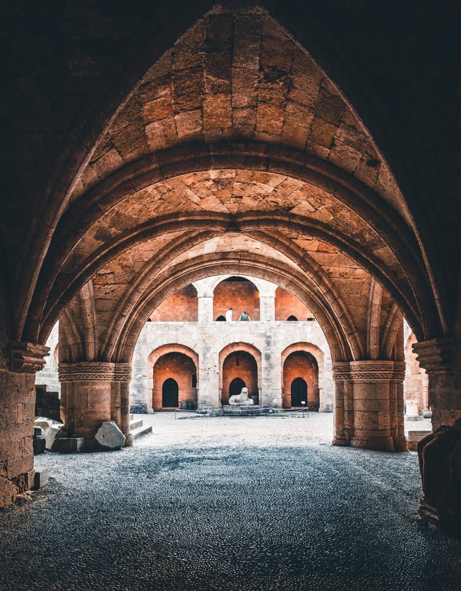 Vue depuis un passage voûté en arches de pierre rouge vers une cour intérieure avec trois arcades blanches et ocre