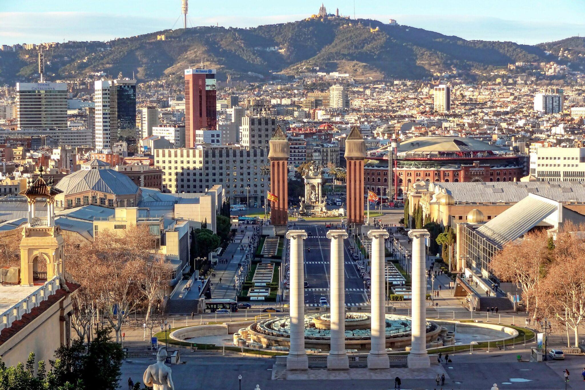 Vue aérienne d’une place avec quatre colonnes monumentales, bâtiments historiques et colline boisée en arrière-plan urbain