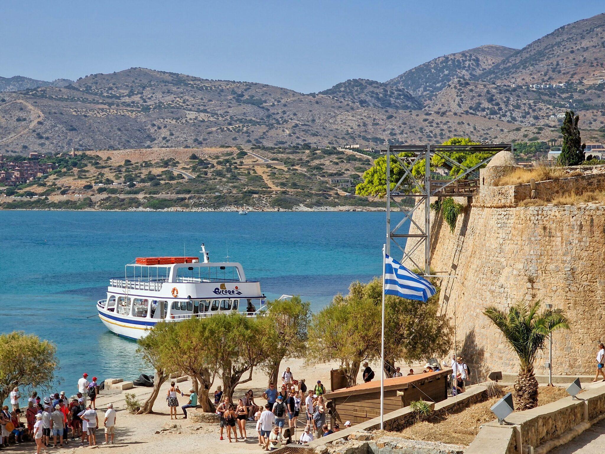 Un bateau touristique blanc et bleu accosté près d’une plage avec drapeau grec et montagne aride en arrière-plan