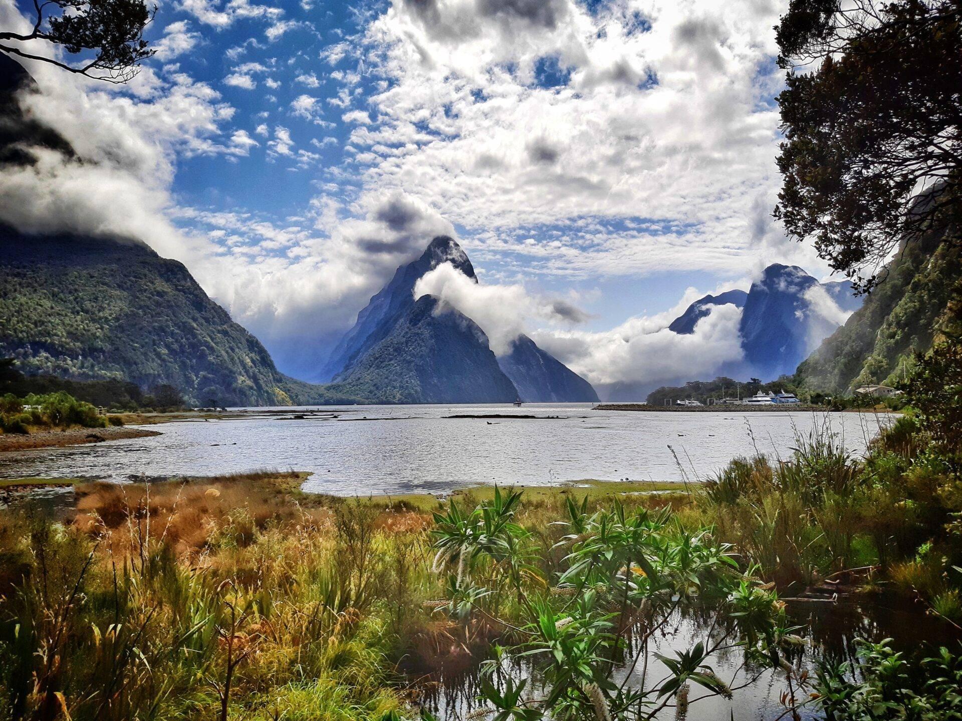 Fjord montagneux avec pics rocheux enveloppés de nuages, lac calme et végétation luxuriante en premier plan