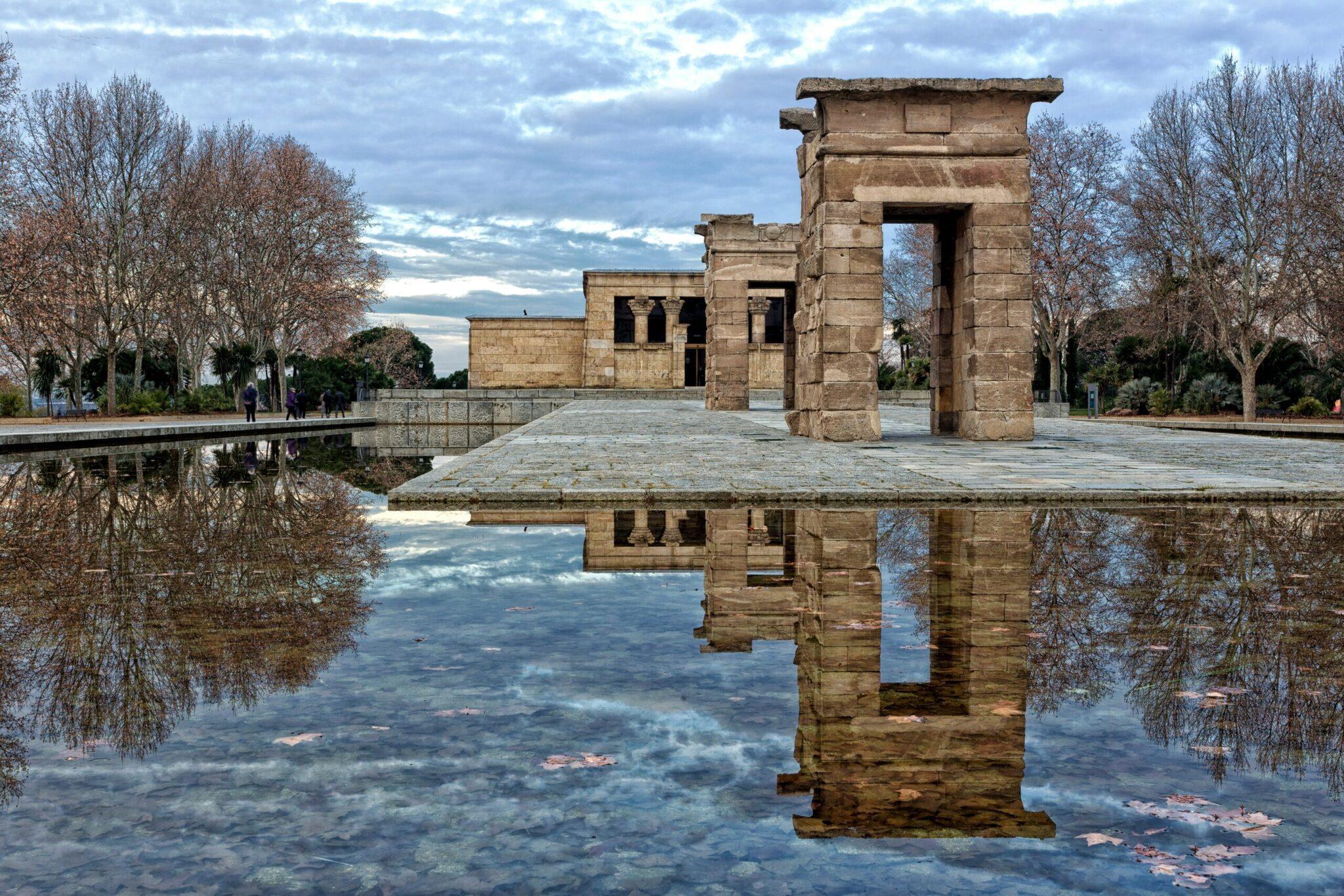 Les ruines d’un temple antique se reflètent dans un plan d’eau calme, entouré d’arbres nus et d’un ciel nuageux