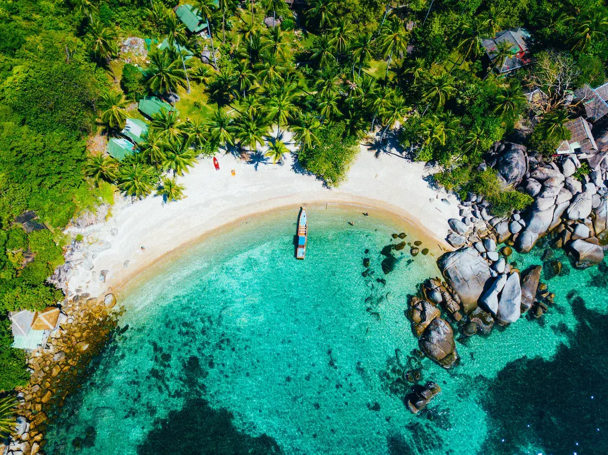 Vue aérienne d’une baie tropicale avec plage de sable blanc, eaux turquoise cristallines, rochers et végétation dense